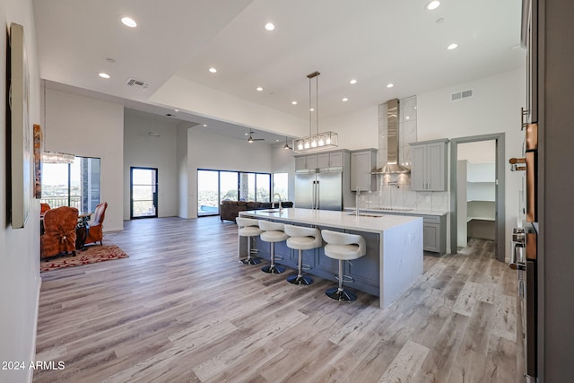 kitchen with wall chimney exhaust hood, a kitchen breakfast bar, a towering ceiling, a kitchen island with sink, and stainless steel built in fridge