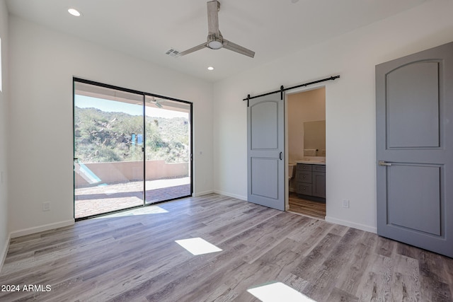 unfurnished bedroom featuring access to exterior, ensuite bath, ceiling fan, a barn door, and light hardwood / wood-style floors