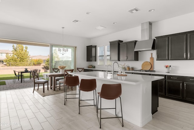 kitchen featuring pendant lighting, wall chimney exhaust hood, light hardwood / wood-style floors, an island with sink, and stainless steel gas stovetop