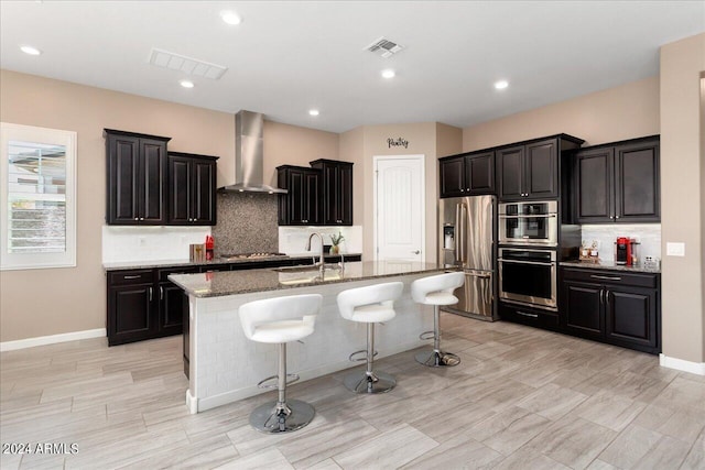 kitchen featuring appliances with stainless steel finishes, a breakfast bar, a kitchen island with sink, sink, and wall chimney range hood