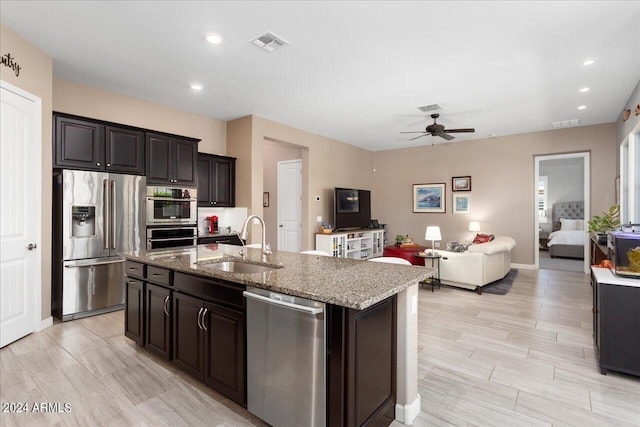 kitchen featuring sink, ceiling fan, an island with sink, light stone counters, and stainless steel appliances