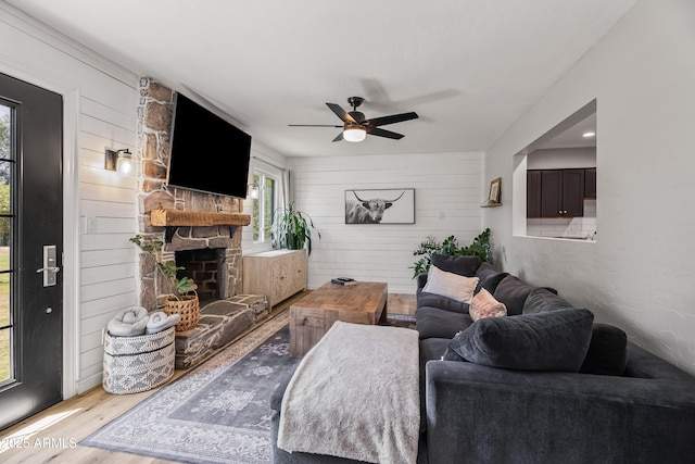 living room featuring wood finished floors, wooden walls, a ceiling fan, and a stone fireplace