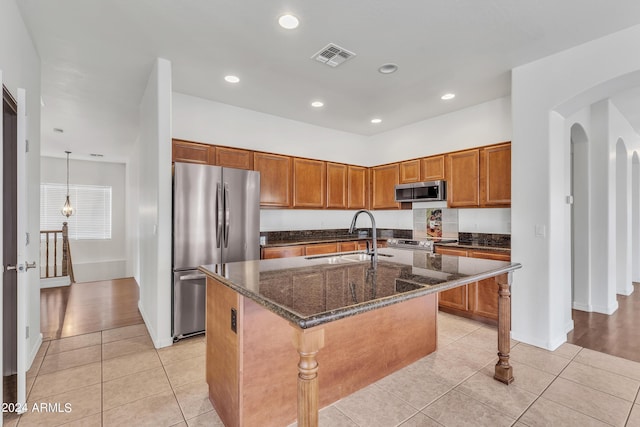 kitchen with a center island with sink, light tile patterned floors, brown cabinets, stainless steel appliances, and a sink