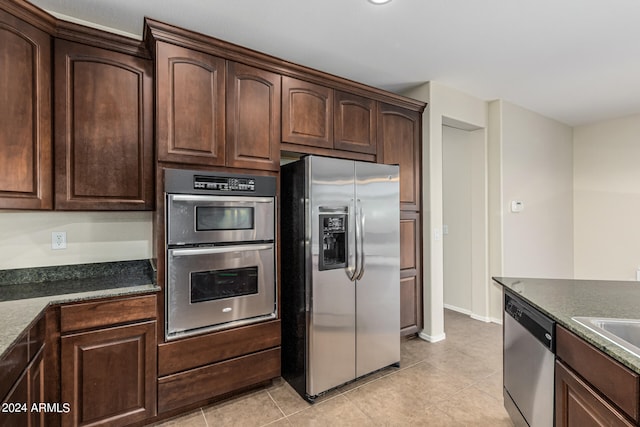 kitchen with dark brown cabinets, appliances with stainless steel finishes, light tile patterned floors, and dark stone counters