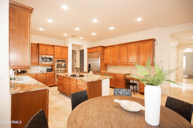 kitchen with light stone counters, light tile patterned floors, a center island with sink, built in appliances, and decorative backsplash