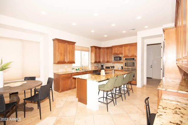 kitchen with light stone counters, tasteful backsplash, a kitchen island with sink, appliances with stainless steel finishes, and a kitchen bar