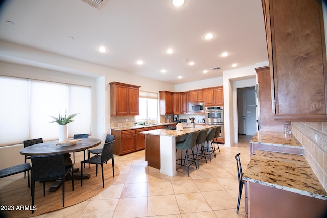 kitchen featuring decorative backsplash, a center island with sink, stainless steel appliances, light stone countertops, and a kitchen bar