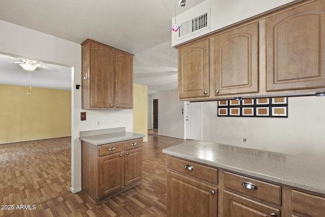 kitchen featuring visible vents, a ceiling fan, light countertops, brown cabinetry, and dark wood finished floors