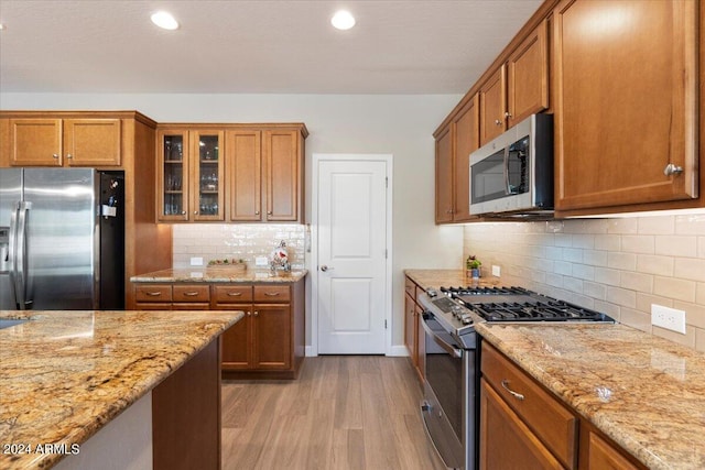 kitchen featuring light wood finished floors, brown cabinetry, appliances with stainless steel finishes, glass insert cabinets, and light stone countertops