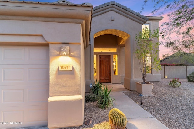 exterior entry at dusk featuring an attached garage, stucco siding, and a tiled roof