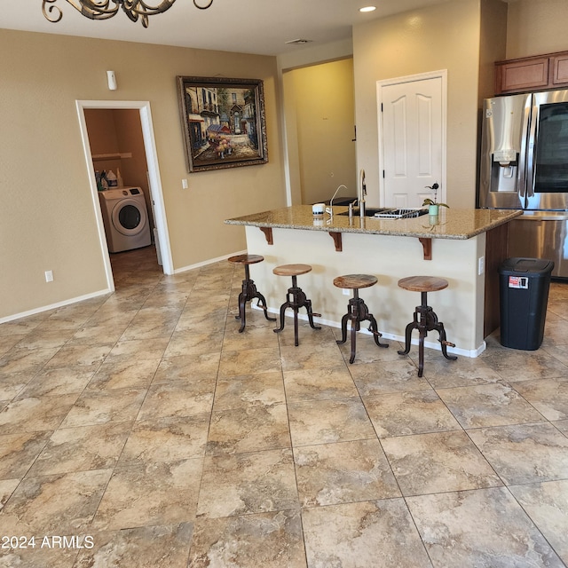 kitchen with light stone countertops, stainless steel refrigerator with ice dispenser, washer / dryer, a kitchen bar, and a kitchen island with sink