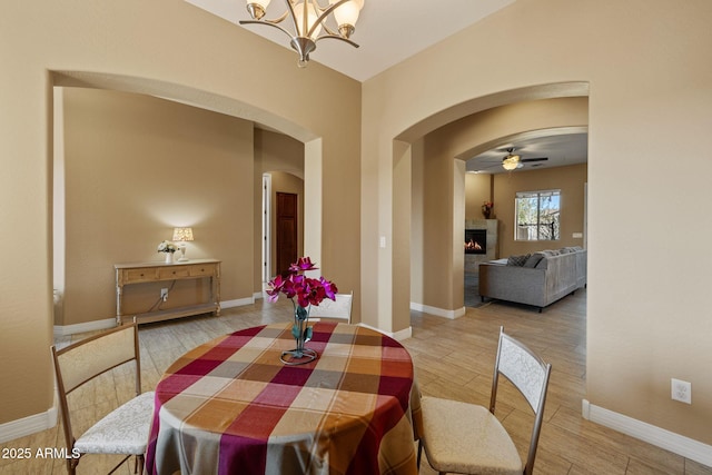dining area with arched walkways, ceiling fan with notable chandelier, baseboards, wood finished floors, and a lit fireplace