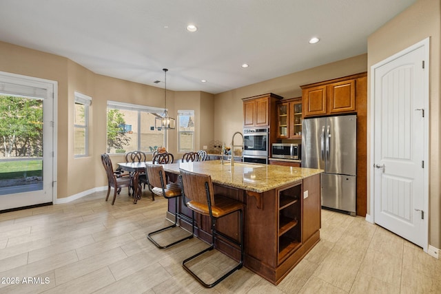 kitchen featuring brown cabinetry, light stone countertops, baseboards, a center island with sink, and stainless steel appliances