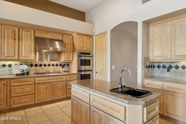 kitchen with arched walkways, under cabinet range hood, a sink, appliances with stainless steel finishes, and an island with sink