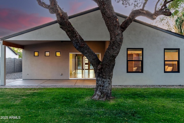 back of house at dusk featuring a yard, a patio, fence, and stucco siding