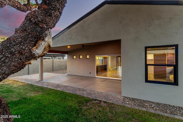 back of house at dusk with a patio, ceiling fan, fence, and stucco siding