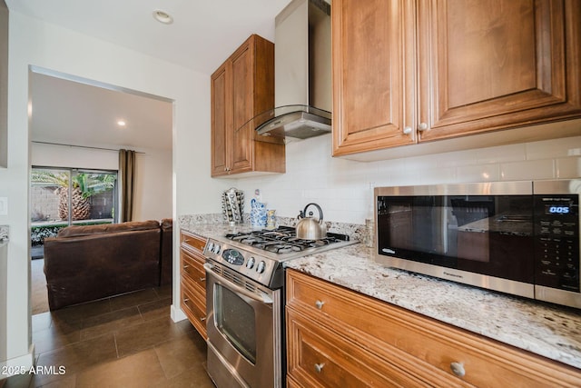 kitchen featuring light stone counters, brown cabinetry, appliances with stainless steel finishes, wall chimney range hood, and backsplash