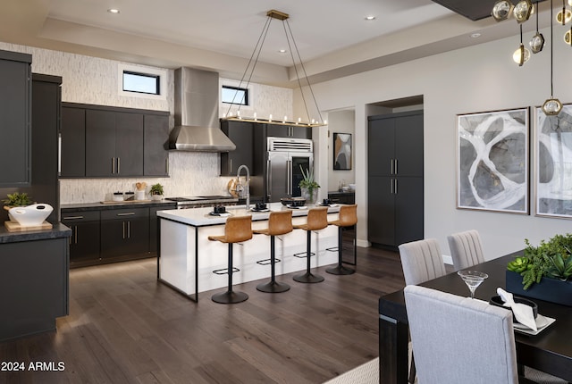 kitchen featuring wall chimney exhaust hood, dark wood-type flooring, a center island with sink, stainless steel built in fridge, and hanging light fixtures