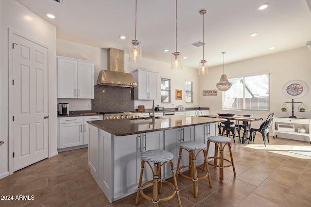 kitchen with a center island with sink, wall chimney exhaust hood, white cabinetry, and hanging light fixtures