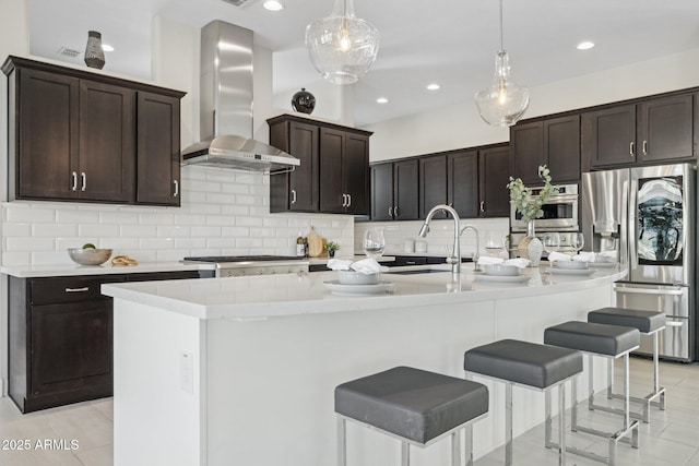 kitchen with light countertops, decorative backsplash, appliances with stainless steel finishes, wall chimney exhaust hood, and a kitchen island with sink