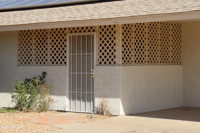 property entrance featuring a shingled roof, a gate, roof mounted solar panels, and stucco siding