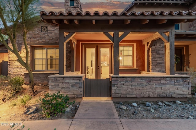 doorway to property featuring covered porch, stone siding, a tiled roof, and a gate