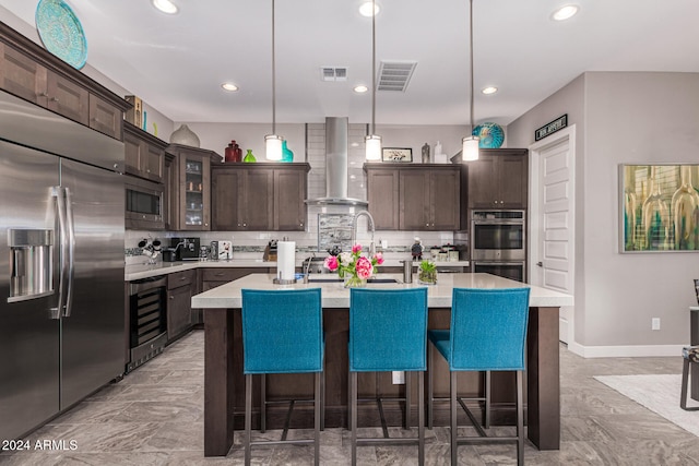 kitchen with built in appliances, a center island with sink, decorative light fixtures, and wall chimney range hood