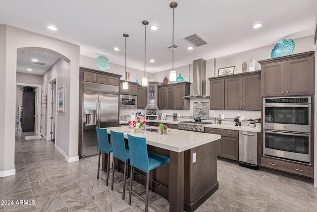 kitchen featuring wall chimney exhaust hood, tasteful backsplash, built in appliances, decorative light fixtures, and dark brown cabinets