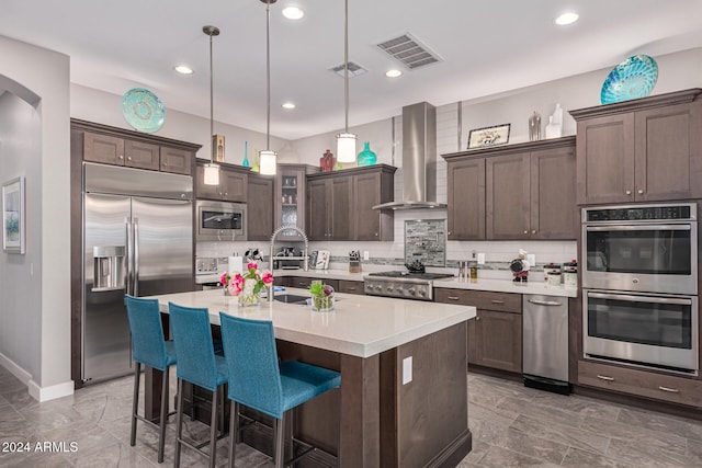 kitchen with dark brown cabinetry, wall chimney range hood, built in appliances, an island with sink, and decorative light fixtures