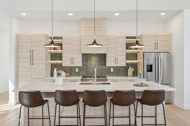 kitchen with light brown cabinetry, decorative light fixtures, stainless steel fridge, decorative backsplash, and a center island with sink