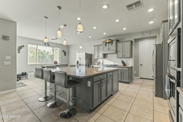 kitchen featuring a center island with sink, gray cabinets, tasteful backsplash, decorative light fixtures, and stainless steel appliances