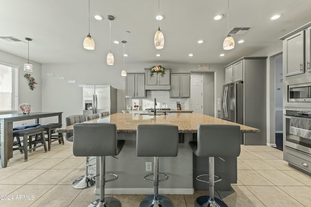 kitchen with gray cabinetry, an island with sink, decorative light fixtures, light stone counters, and stainless steel appliances