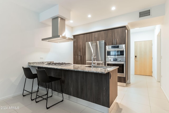 kitchen with dark brown cabinetry, light stone countertops, island exhaust hood, kitchen peninsula, and appliances with stainless steel finishes