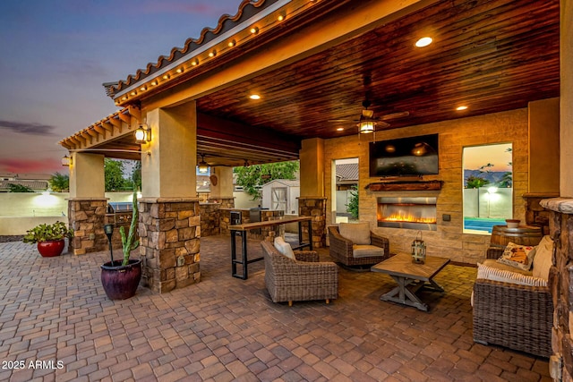 patio terrace at dusk with a storage shed, an outdoor living space with a fireplace, a ceiling fan, and an outdoor structure