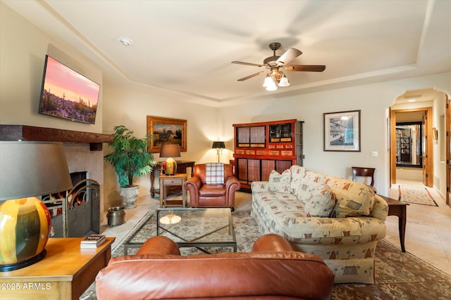 tiled living room featuring a tray ceiling, a ceiling fan, arched walkways, and a warm lit fireplace