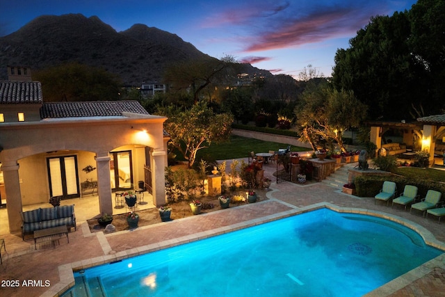 pool at dusk with an outdoor pool, a mountain view, a patio, and a warm lit fireplace