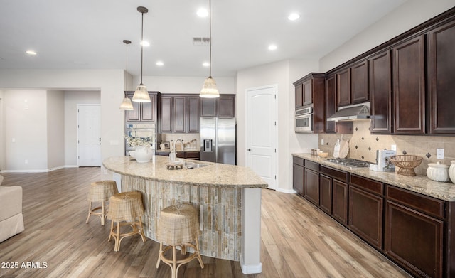 kitchen featuring stainless steel appliances, an island with sink, light wood-type flooring, sink, and decorative light fixtures