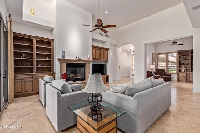 8689 E Krail St, Scottsdale, AZ 85250 - living room featuring a ceiling fan, arched walkways, light tile patterned floors, a towering ceiling, and a glass covered fireplace