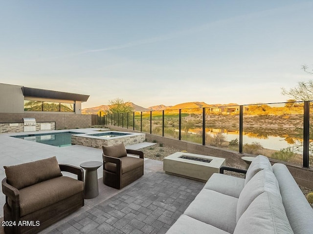 pool at dusk with exterior kitchen, a fire pit, an in ground hot tub, a patio, and a water and mountain view