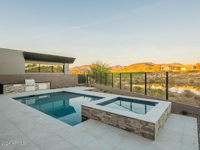 pool at dusk with a patio area, area for grilling, a grill, a mountain view, and an in ground hot tub