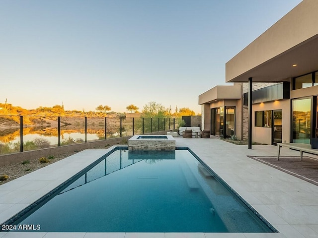 pool at dusk with an in ground hot tub, a patio, and a water view