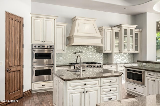 kitchen featuring premium range hood, stainless steel double oven, tasteful backsplash, a sink, and dark wood finished floors