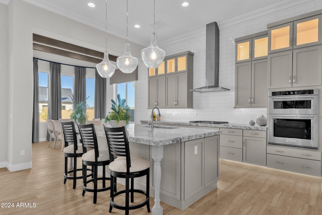 kitchen with stainless steel appliances, hanging light fixtures, sink, wall chimney exhaust hood, and light hardwood / wood-style flooring