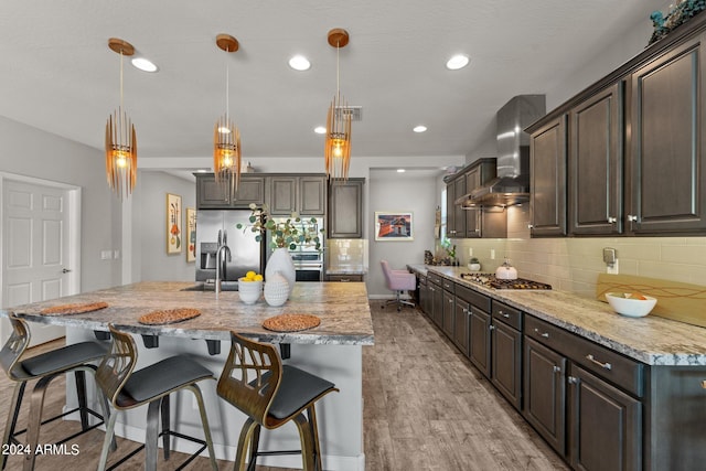 kitchen featuring light wood-type flooring, decorative light fixtures, dark brown cabinetry, and wall chimney range hood