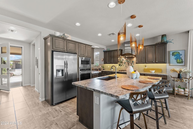 kitchen featuring pendant lighting, sink, wall chimney range hood, stainless steel appliances, and a center island with sink