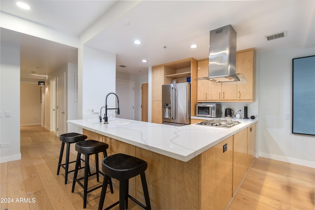 kitchen with light brown cabinets, wall chimney exhaust hood, kitchen peninsula, stainless steel appliances, and light wood-type flooring