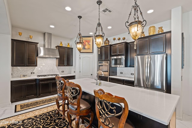 kitchen with a center island with sink, sink, wall chimney exhaust hood, decorative light fixtures, and stainless steel appliances