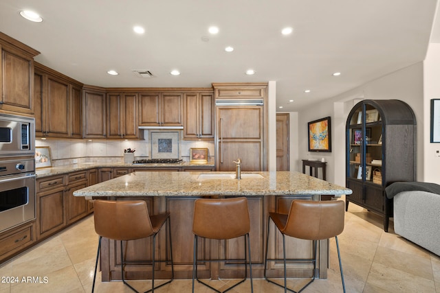 kitchen with built in appliances, a center island with sink, light tile flooring, light stone counters, and a breakfast bar