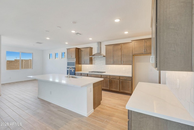 kitchen featuring brown cabinets, a sink, decorative backsplash, wall chimney range hood, and light wood-type flooring