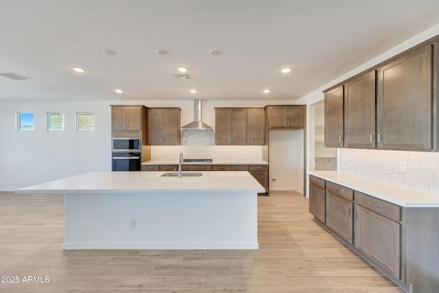 kitchen with an island with sink, a sink, stainless steel appliances, wall chimney range hood, and wood tiled floor